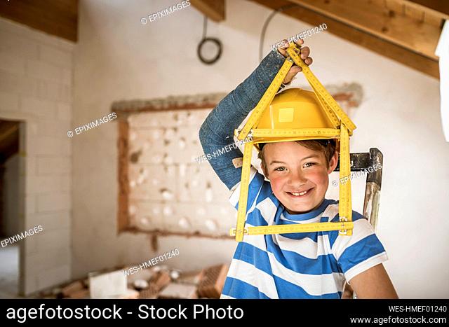 Smiling boy holding tape measure of house shape at attic during renovation
