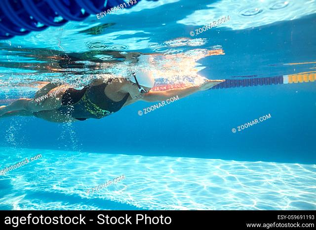 Athletic female swimmer swims underwater in the swimming pool outdoors. She wears a black-gray swimsuit with patterns, a white swim cap and swim glasses