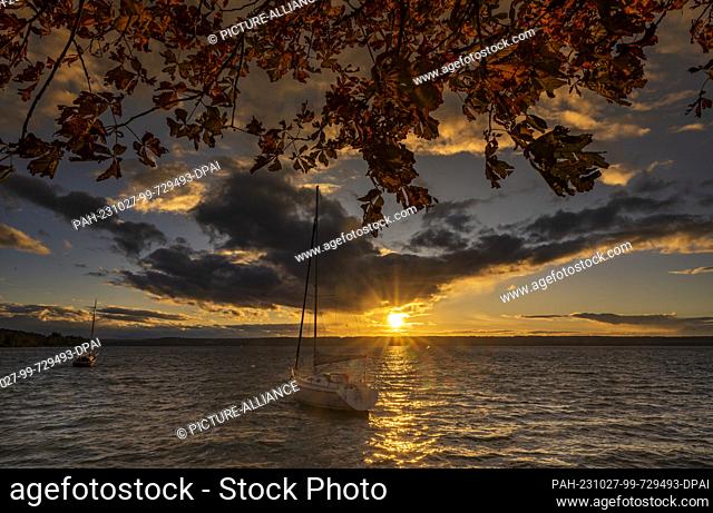 27 October 2023, Bavaria, Herrsching: The sun is over the western shore of the Ammersee in the evening, where sailing ships are anchored