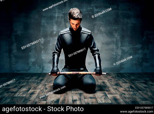 Warrior man sitting on floor posing with a sword over black background