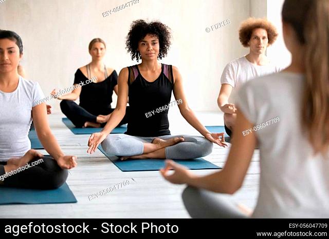 Female coach training diverse young people rear view, practicing yoga at group lesson, doing Padmasana exercise, sitting in Lotus pose on mats, working out