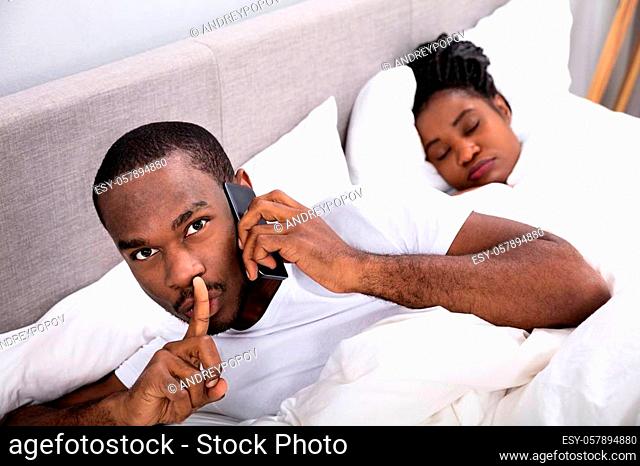 Close-up Of A Young African Man Talking Secretly On Cellphone While His Wife Sleeping