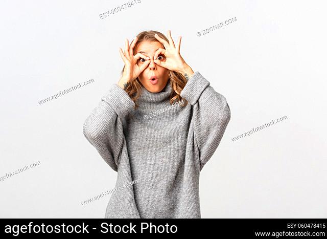 Portrait of funny blond girl making faces, showing finger glasses and mocking someone, standing over white background