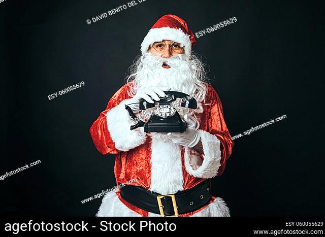 Man dressed as Santa Claus holding a retro telephone from the 60's, on black background. Christmas concept, Santa Claus, gifts, celebration