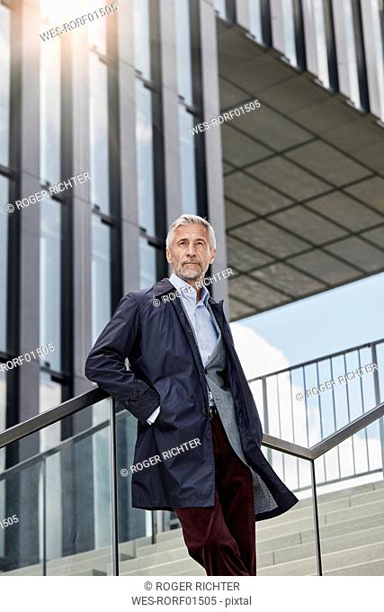 Portrait of mature businessman standing on stairs in front of modern office building looking at distance