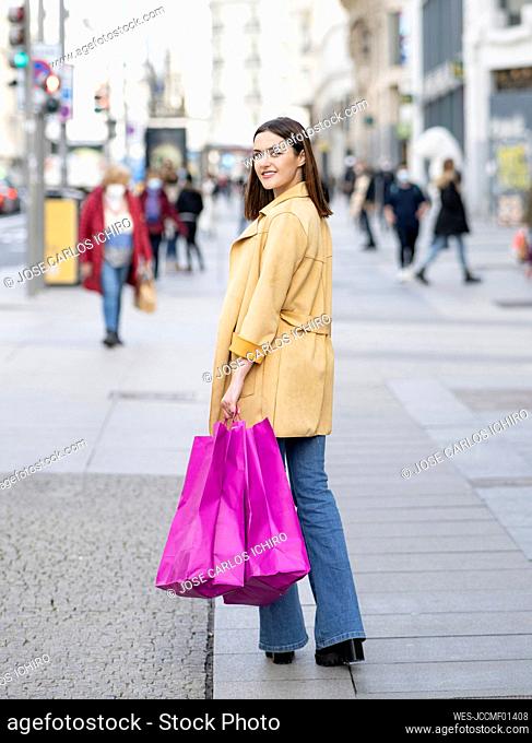 Smiling young woman with magenta colored shopping bags walking on footpath in city