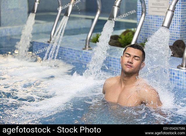 Horizontal indoors shot of handsome man with eyes closed resting under stream of shower in pool