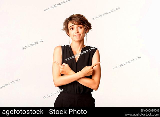 Clueless silly girl smiling and pointing fingers sideways, asking for help with choice, making decision, standing over white background