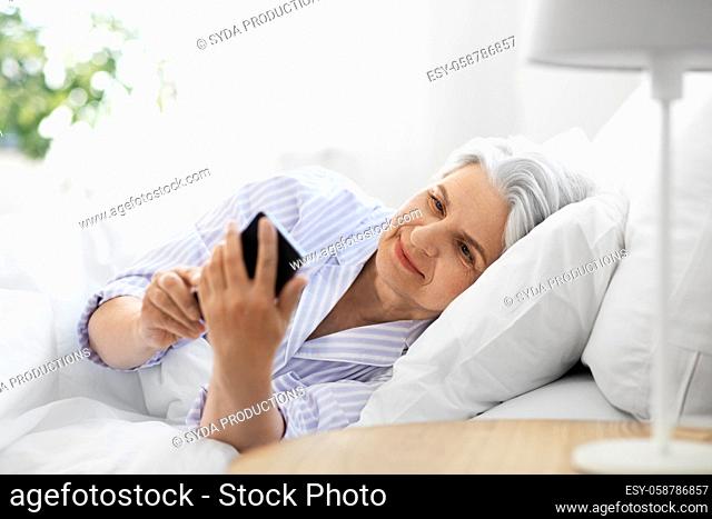happy senior woman with smartphone in bed at home