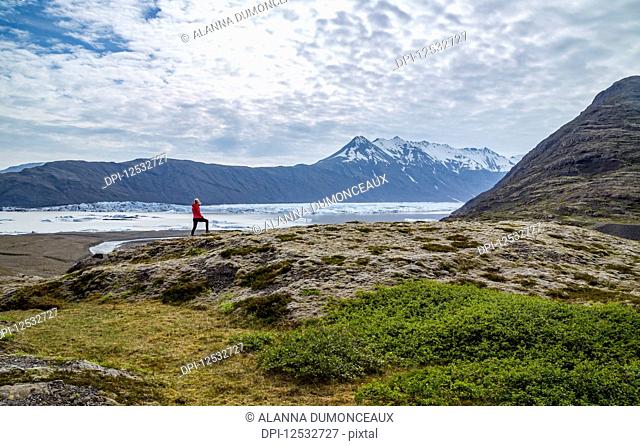 A female hiker walks along the edge of a hill on the trail above a glacier lake and mountain viewpoint in Southern Iceland, Vatnajokull National Park; Iceland