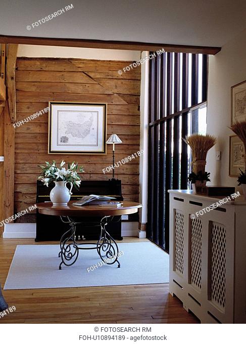 Circular wood table with metal legs in barn conversion hall with wood panelled wall and white fretwork radiator cover