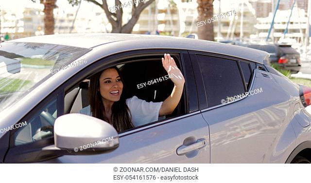 Beautiful young woman sitting inside of modern car and waving with hand greeting someone in sunny day at street