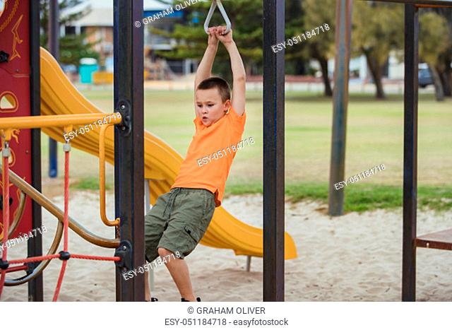 A side-view shot of a young caucasian boy wearing casual clothing, he is swinging from a metal frame in a public park