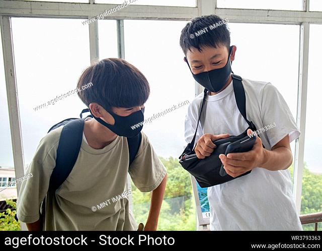 Two 13 year old Japanese boys in face masks, friends looking at a mobile phone screen
