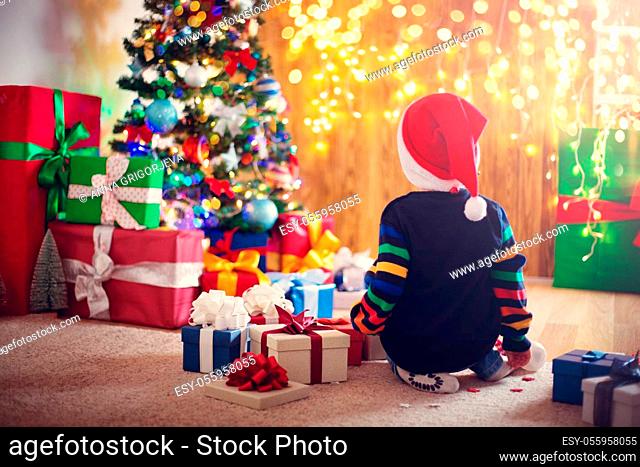 Boy and sitting on the floor with presents near christmas tree. Child in red hat at home in winter