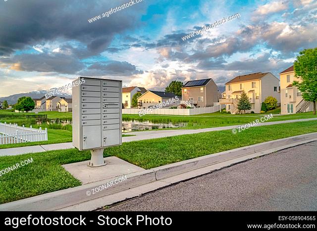 Mailbox on the side of the road with pond grassy terrain and homes background. Snow capped mountain and cloudy sky can also be seen in this landscape