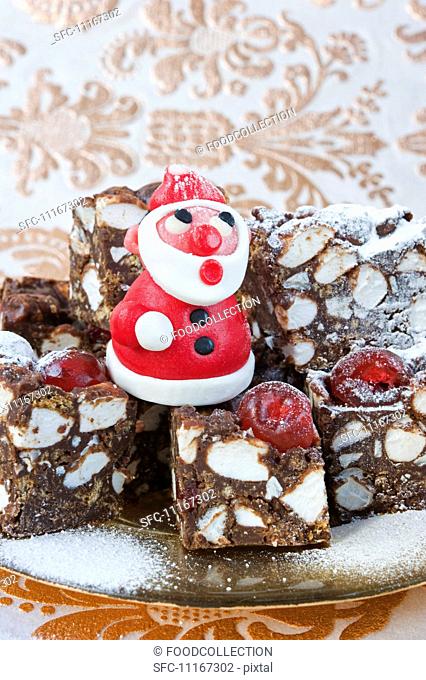 a model in red marzipan of father Christmas standing on squares of rocky -road cakes for Christmas with gold patterened paper