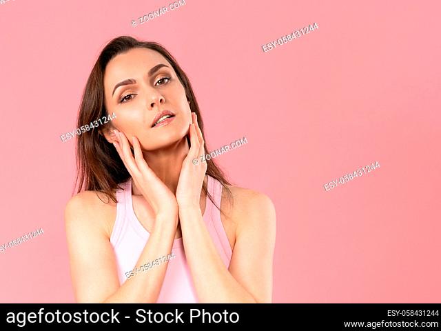Moisturising her face beautiful woman with no makeup standing with hands on her chicks, massaging it gently attractive brunette girl on pink background