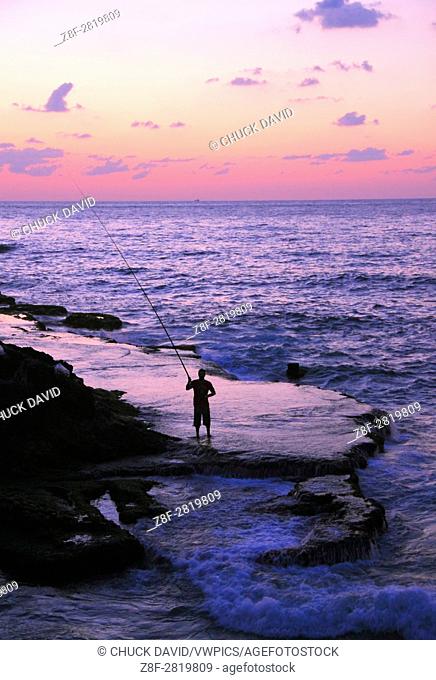 A lone fisherman casts his rod along Beirut's rocky Mediterranean shoreline at sunset, Lebanon