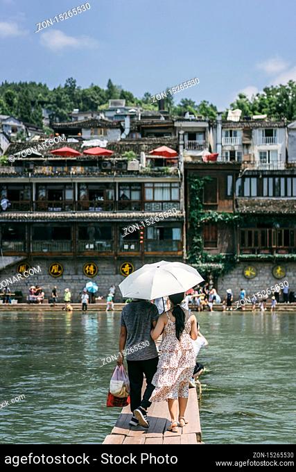 Feng Huang, China - August 2019 : Couple holding umbrella crossing waters on stepping stones on Tuojiang river, flowing through the centre of Fenghuang Old Town