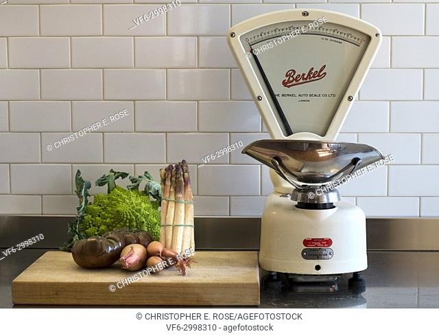 A few choice vegetables wait on a chopping board on a stainless steel kitchen work surface alongside a set of classic vintage grocers weighing scales made by...