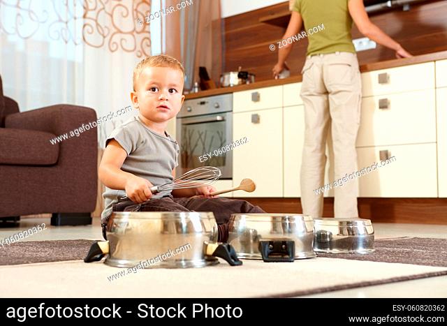 Little boy sitting on carpet in kitchen playing with cooking pots, mother preparing food in background