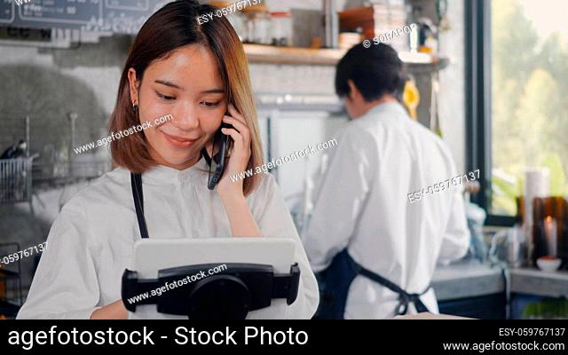 Happy Asian barista woman with apron coffee maker receive phone call and record pre order to computer in coffee cafe shop
