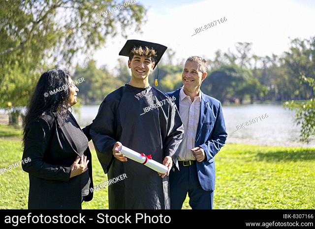 Young recently graduated boy, dressed in cap and gown, with his degree in hands, celebrating with his multi ethnic family on the university campus