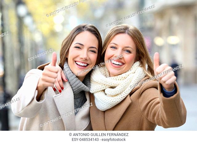 Front view portrait of two friends wearing coats smiling at camera with thumbs up in winter on the street