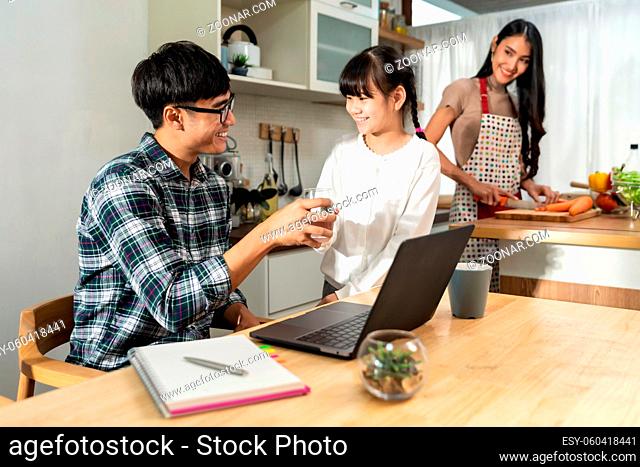 Asian little girl serving water to her dad while working with computer laptop and mom cooking in kitchen. Family Work at home while quarantine from COVID-19