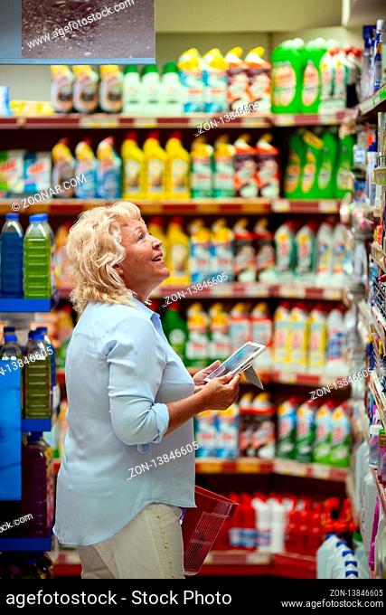 A smiling middle aged woman in a light blue shirt is standing in a household section of a supermarket. She is holding a tablet and a red shopping basket in her...