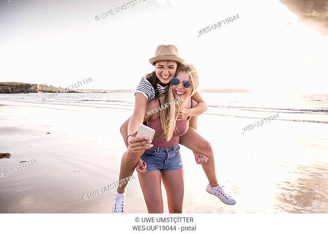 Two girlfriends having fun on the beach, carrying each other piggyback, taking smartphone selfies