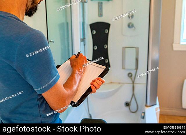 A close up and rear view of a man using a notepad and pen during a home inspection inside a bathroom, with blurry shower cubicle in background and copy-space
