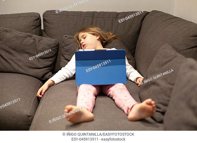five years old blonde child in pajamas sleeping lying on the coach, with digital tablet on her legs