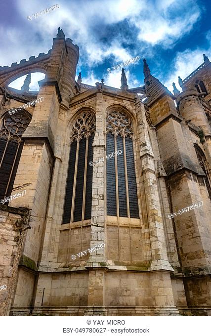 Gothic Cathedral of Narbonne seen from ground with dark architectural structures against sky and clouds, city in the south of France