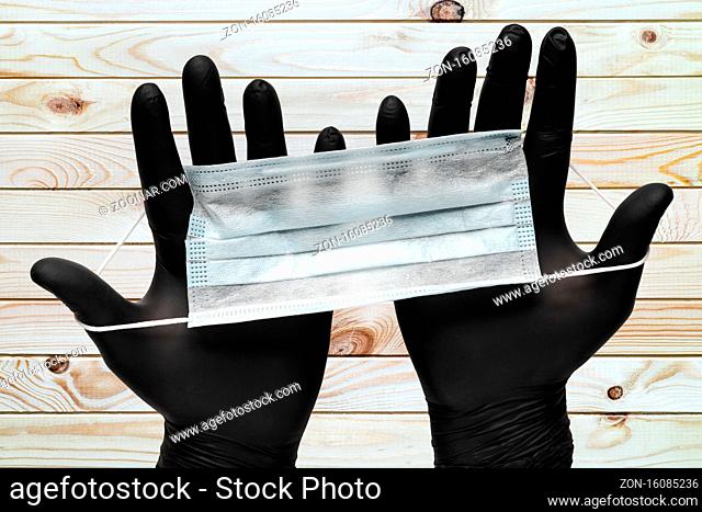 Doctor holds surgical face masks in two hands in black medical gloves on wooden plank brown background. Concept pandemic outbreak, virus prevention