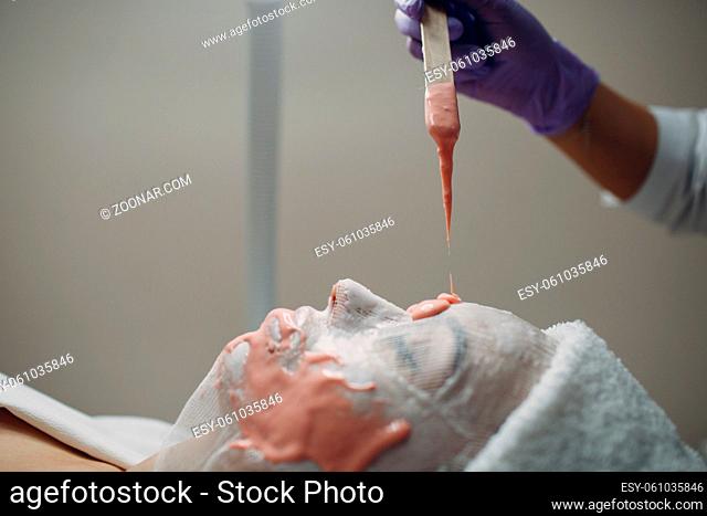 Alginate face mask application process. A female hands of a beautician holding spatula. Woman lying on a couch in office of cosmetologist