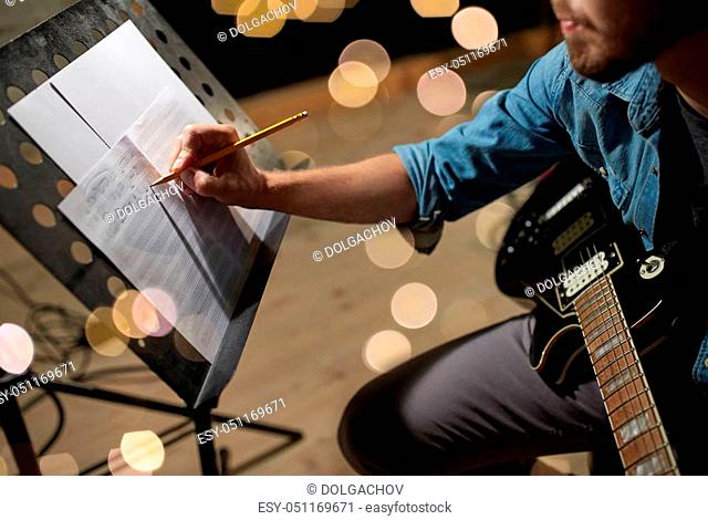 art and creativity concept - man with guitar writing notes to music book at studio over festive lights