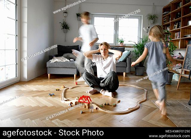 Stressed out mother sitting in the middle of toys, while children are running around her