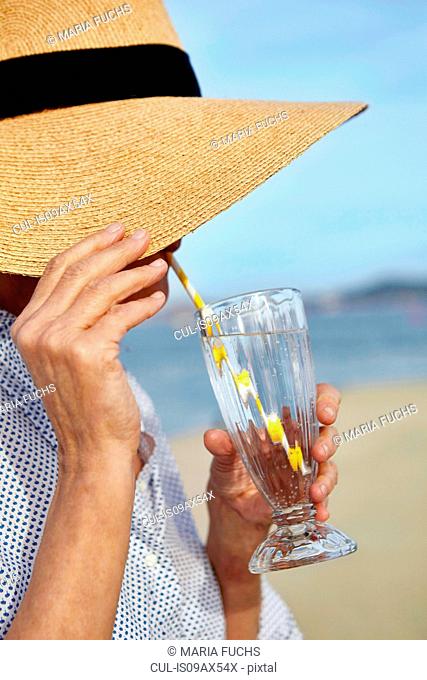 Mature woman wearing straw hat, beside beach, drinking cool drink through drinking straw