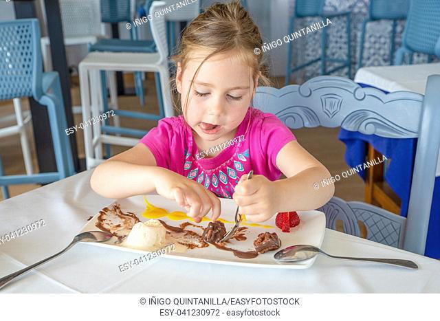funny cute four years old blonde girl eating, and sticking out tongue, with spoon sweet dessert: black chocolate, vanilla ice cream