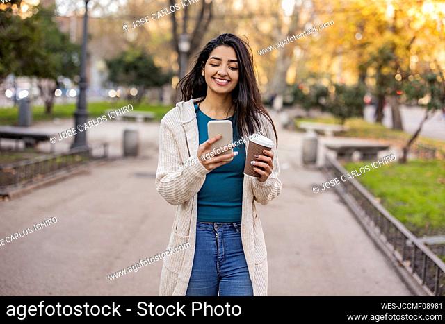 Happy woman with disposable cup using smart phone in park