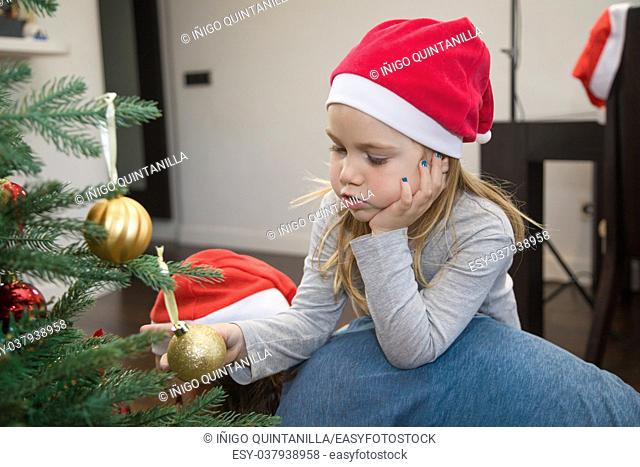 funny scene: four years blonde cute girl with red Santa Claus hat, holding a golden ball in Christmas tree, resting on her mother back, at home
