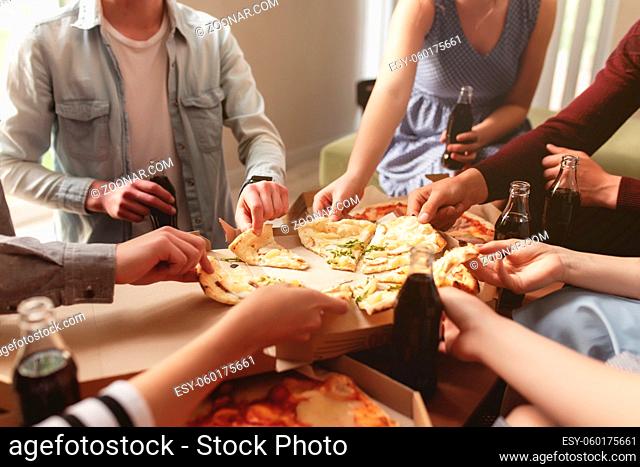 A pizza party, a group of friends chatting and happily eating pizza at the table and drinking sweet soda water. Many friends handing out together because of...