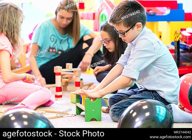 Cute pre-school boy cooperating with his colleagues at the construction of a structure made of wooden toy blocks, under the guidance of a young kindergarten...