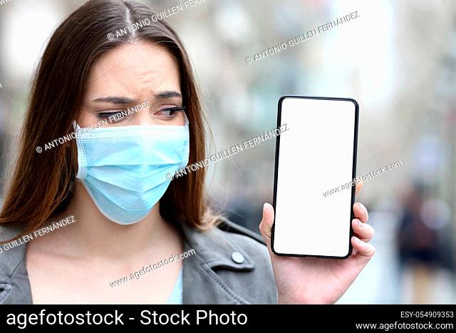 Front view portrait of a scared girl with protective mask preventing contagion looking at her blank smart phone screen