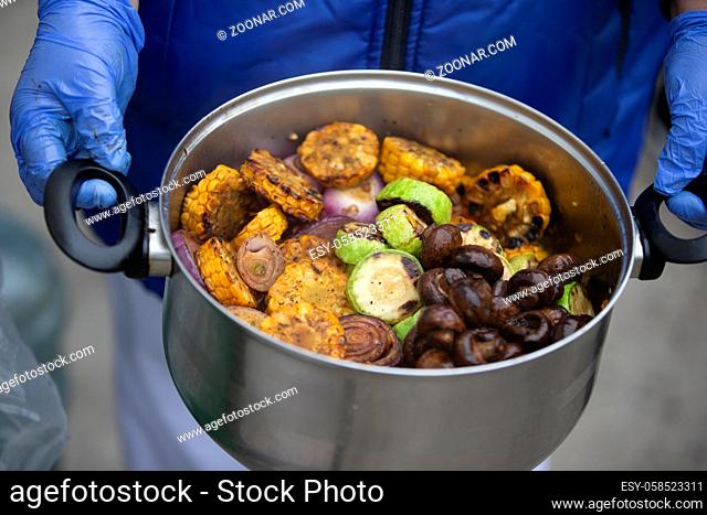 The chef's hands are holding a saucepan with grilled vegetables and mushrooms