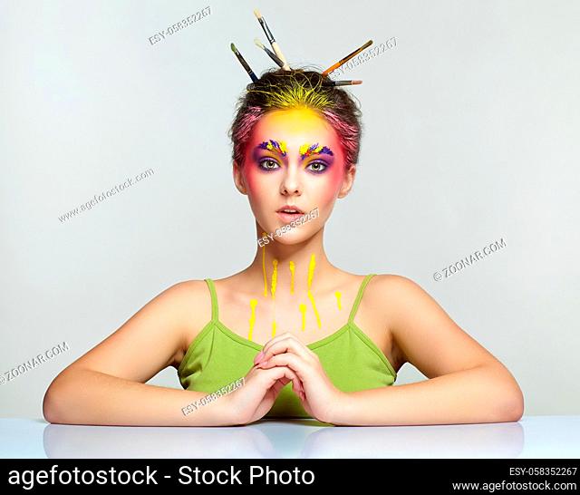 Portrait of young woman posing at the table with brushes in hair. Unusual female art make-up with paint on brows, hair and around eyes