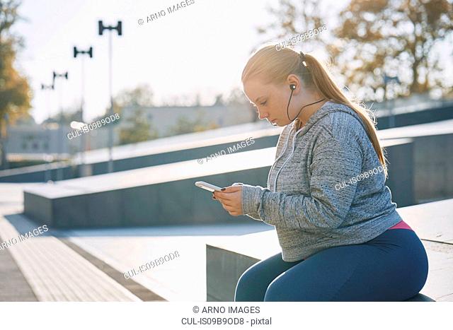 Curvaceous young woman training, sitting on wall looking at smartphone
