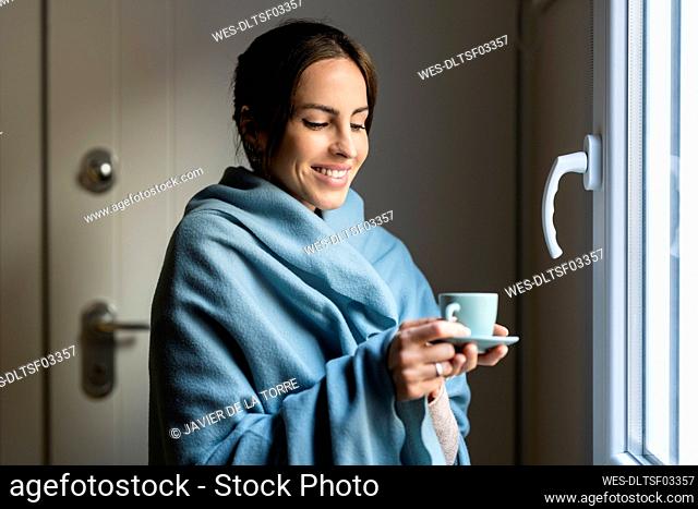 Happy woman wrapped in blanket holding cup of coffee near window